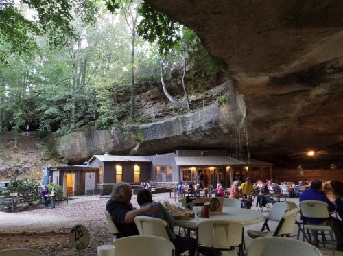 A rustic dining area under a large rock overhang, surrounded by trees and people enjoying their meals.