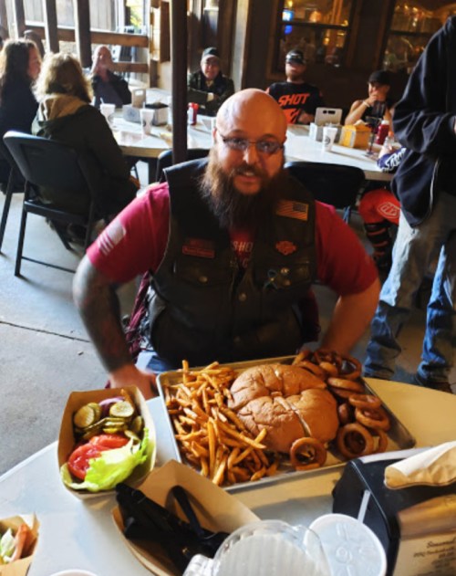 A man with a beard in a motorcycle vest sits at a table with a large burger, fries, and onion rings.