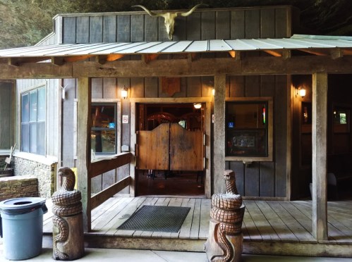 A rustic wooden building with a cow skull above the entrance and carved snake sculptures flanking the door.