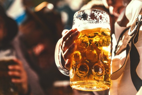A person holding a large beer mug filled with golden beer, with blurred figures in the background.