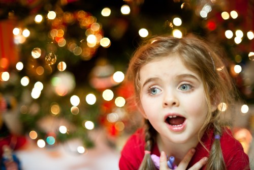 A young girl with pigtails, wearing a red shirt, looks surprised in front of a decorated Christmas tree with blurred lights.