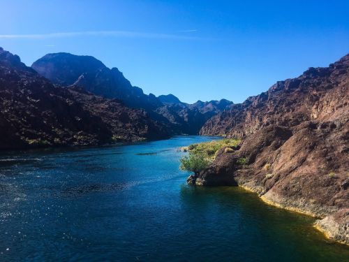 A serene river flows through rocky mountains under a clear blue sky, with greenery along the water's edge.