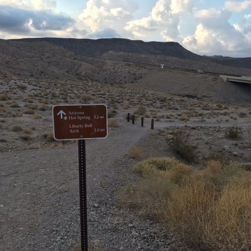 A signpost indicating directions to Arizona Hot Spring and Liberty Bell Arch, with a desert landscape in the background.