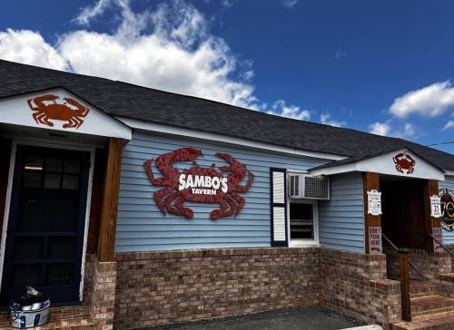 Exterior of Sambo's Tavern featuring a blue facade, brick accents, and crab-themed signage under a cloudy sky.