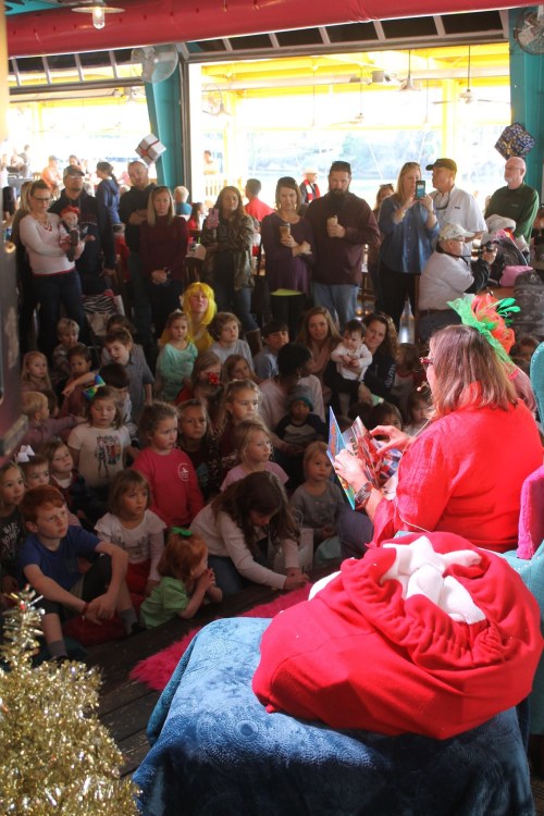 A storyteller in festive attire reads to a large group of captivated children in a colorful indoor setting.