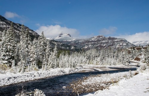 A serene winter landscape featuring a snow-covered river, trees, and mountains under a clear blue sky.