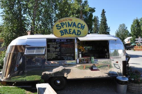 A shiny silver food truck named "Talkeetna Spinach Bread" with a menu board and surrounded by trees.