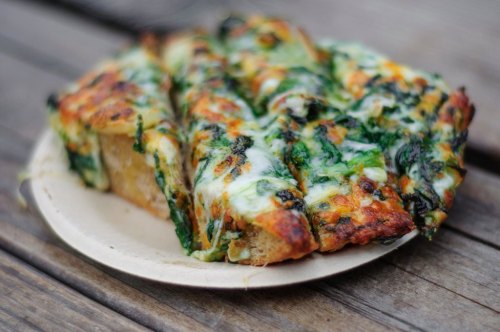 A slice of cheesy spinach pizza on a paper plate, resting on a wooden table.