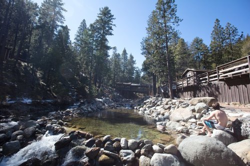 A boy sits on rocks by a clear stream surrounded by tall pine trees and wooden structures under a bright blue sky.