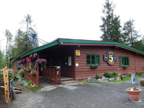 A rustic wooden building with green trim, satellite dish, and flower pots, surrounded by trees and gravel path.