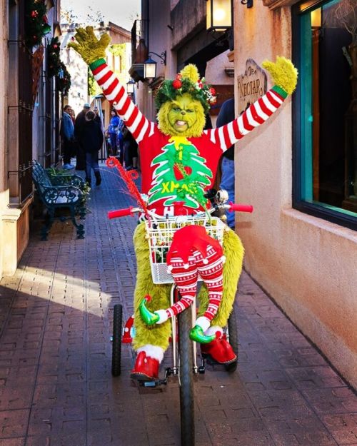 A festive Grinch character on a tricycle, wearing a Christmas sweater and surrounded by holiday decorations.