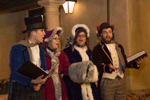 A group of four carolers in festive attire singing joyfully outdoors, surrounded by warm lighting.