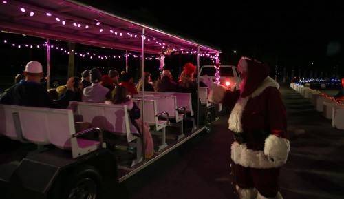 A festive scene with a Santa figure waving to a decorated tram filled with cheerful passengers at night.