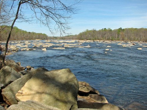 A serene river scene with large rocks and trees lining the banks under a clear blue sky.
