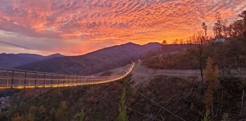 A suspension bridge stretches across a valley under a vibrant sunset with colorful clouds and mountains in the background.