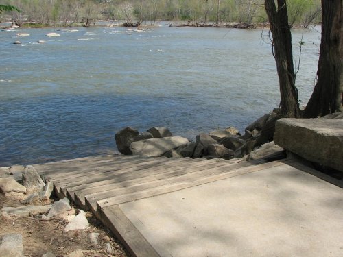 A wooden path leads down to a riverbank, surrounded by rocks and trees, with calm water flowing nearby.