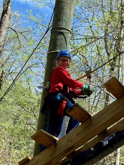 A person in a helmet and harness smiles while climbing a wooden structure in a forested area.