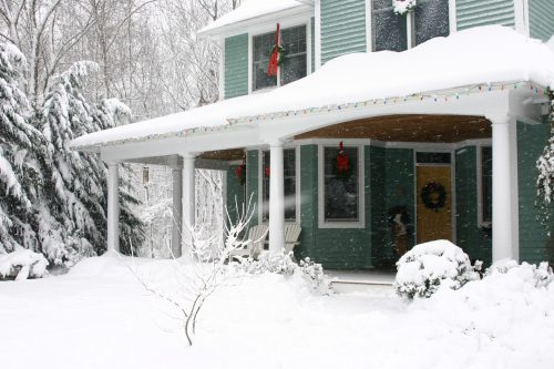 A snow-covered house with a porch, decorated with wreaths and lights, surrounded by trees in a winter landscape.