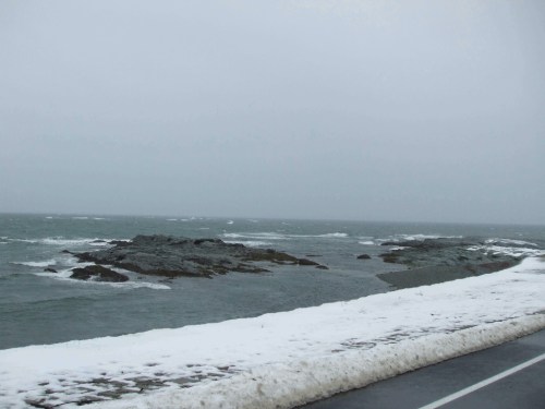 A gray, overcast day at the coast, with rocky shores and patches of snow along the water's edge. Waves gently crash.