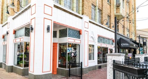 A corner building with large windows, featuring a mix of white and orange accents, surrounded by a brick sidewalk.