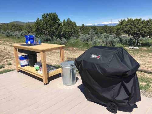A black grill cover on a patio, with a wooden table, trash can, and cooler nearby, set against a scenic outdoor backdrop.