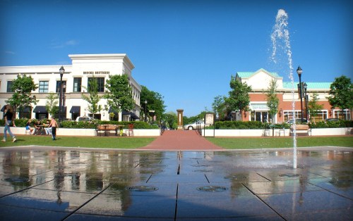 A sunny plaza with a fountain, surrounded by trees and buildings, featuring people walking and benches for seating.