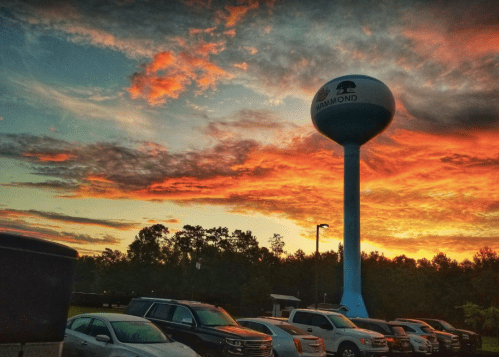 A water tower stands against a vibrant sunset, with colorful clouds and parked cars in the foreground.