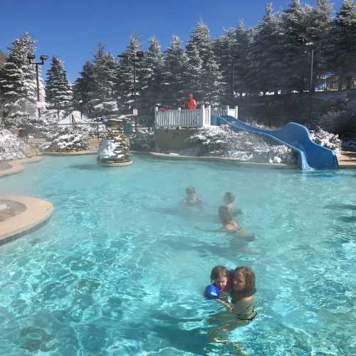 A snowy landscape surrounds a steaming pool where children play, with a water slide and a person on a deck in the background.