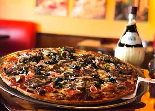 A large pizza topped with meats, olives, and vegetables, next to a bottle of olive oil on a wooden table.