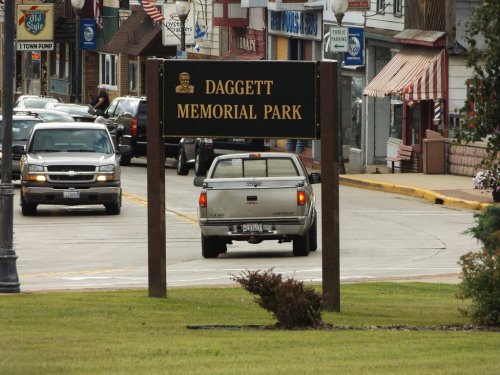 Sign for Daggett Memorial Park with vehicles on a street and shops in the background. Green grass in the foreground.