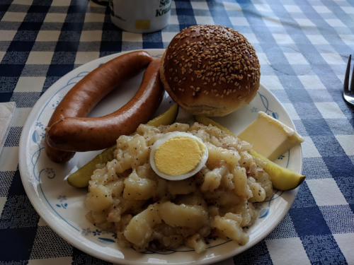 A plate with a sausage, a sesame seed bun, potato salad with an egg, and pickles on a checkered tablecloth.