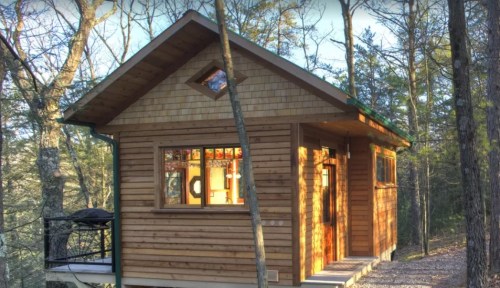 A cozy wooden cabin nestled among trees, featuring large windows and a sloped roof.