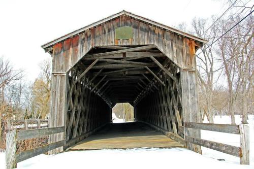A rustic wooden covered bridge with a snow-covered landscape, showcasing its intricate lattice design and open entrance.