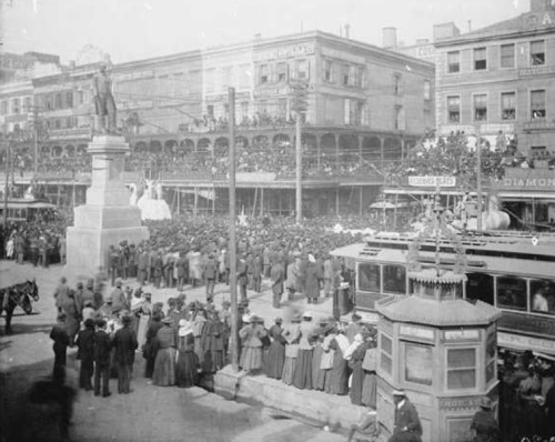 A historic crowd gathers around a statue, with buildings and a streetcar in the foreground, capturing a lively public event.
