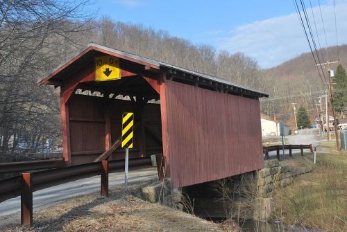 A red covered bridge spans a small stream, with a warning sign indicating a height limit. Trees and a road are nearby.