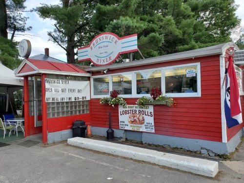 A red diner with a sign reading "Brunswick Diner," surrounded by greenery and flowers, open daily for meals.
