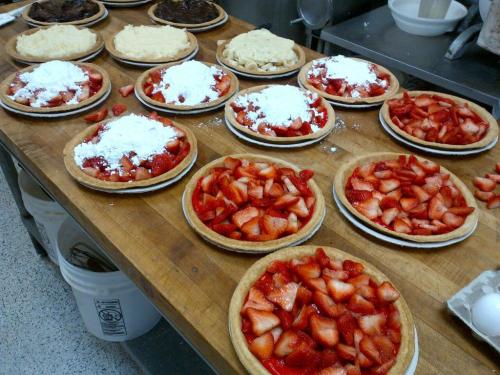 A wooden table with several fruit pies topped with strawberries and powdered sugar, alongside unbaked pie crusts.