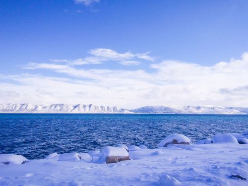 A serene winter landscape featuring a snowy shoreline and calm blue waters under a clear sky with distant mountains.