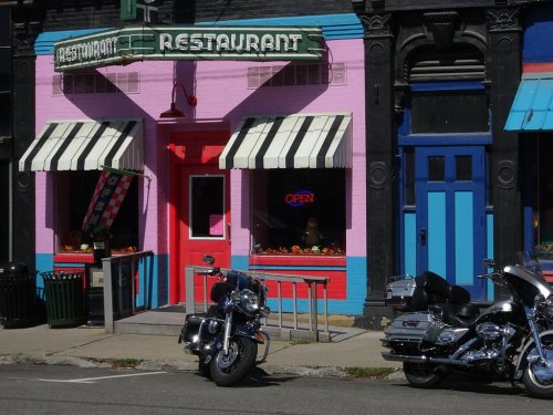 Colorful restaurant exterior with pink walls, striped awnings, and two parked motorcycles in front.