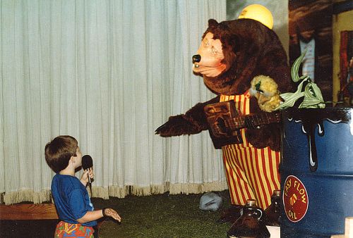 A child with a microphone interacts with a large bear character holding a guitar in a colorful, playful setting.