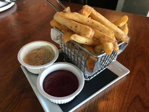 A basket of golden fries served with two dipping sauces: ketchup and a seasoned spice mix on a wooden table.