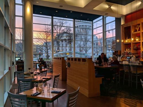 A cozy restaurant interior with large windows showcasing a sunset and a view of a nearby building.
