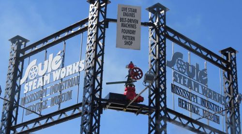 Sign for Joule Steam Works featuring steam engines, a foundry, and a pattern shop against a blue sky.