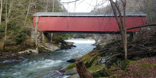 A red covered bridge spans a river, surrounded by trees and rocky banks, with a waterfall in the background.