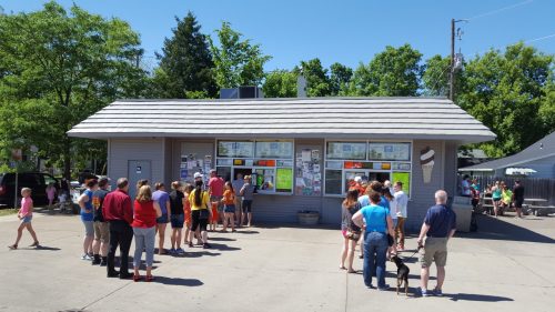 A crowd gathers outside a small building with large windows displaying colorful posters on a sunny day.