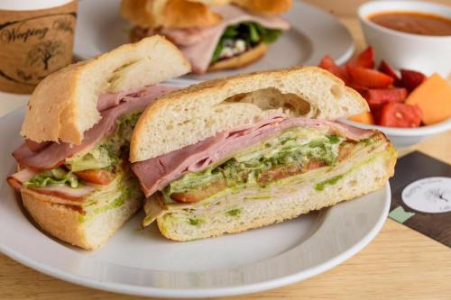 A sandwich with layers of turkey, ham, lettuce, and tomato on a plate, accompanied by fruit and soup in the background.