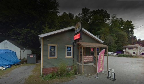 A small green building with a sign, featuring a porch and a pink banner, surrounded by trees and parked vehicles.