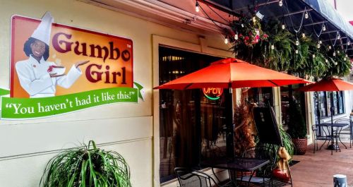 Sign for "Gumbo Girl" restaurant featuring a chef and the slogan, "You haven't had it like this!" with outdoor seating.