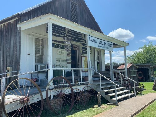 A rustic store with a porch, featuring signs for drinks and crafts, surrounded by greenery and old wagon wheels.