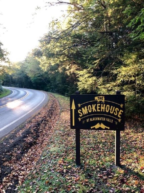 A sign for "The Smokehouse at Blackwater Falls" beside a winding road, surrounded by trees and autumn foliage.